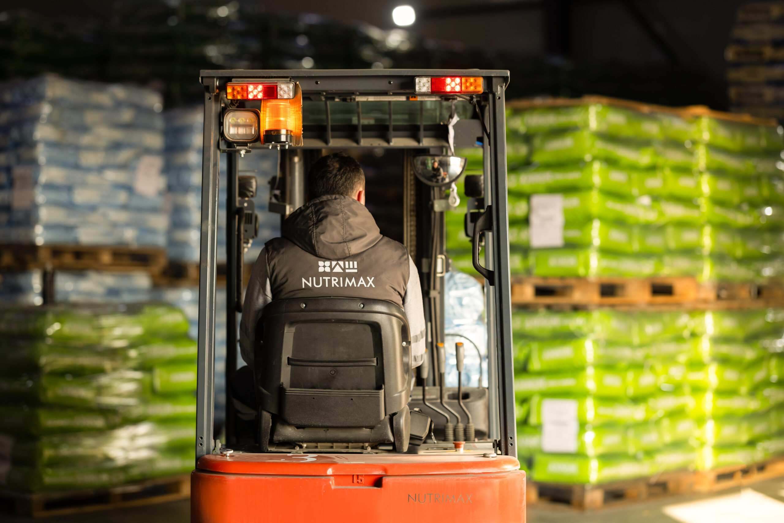 Nutrimax worker driving a forklift in a warehouse filled with stacked green and blue bags